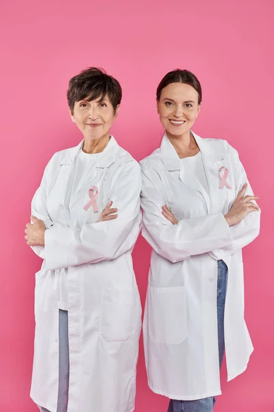Smiling female doctors with ribbons crossing arms isolated on pink, breast cancer awareness — Stock Photo