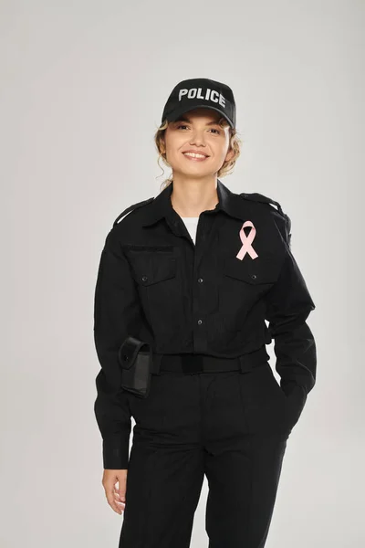 Poliziotta sorridente con nastro rosa in posa isolata su grigio, consapevolezza del cancro al seno — Foto stock