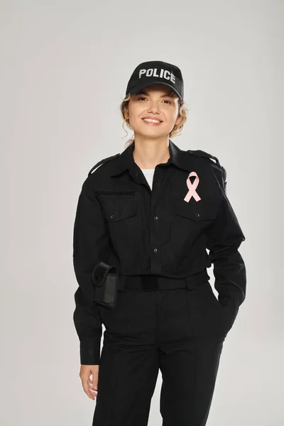 Mujer policía sonriente con cinta rosa posando aislado en gris, conciencia de cáncer de mama - foto de stock