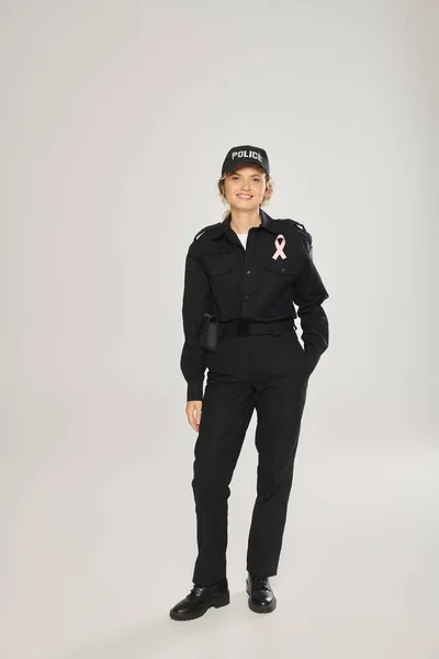 Full length of smiling policewoman with pink ribbon on uniform on grey background, breast cancer — Stock Photo