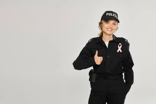 Lächelnde Polizistin mit rosafarbener Schleife, Daumen hoch auf grau, Brustkrebsmonat — Stockfoto