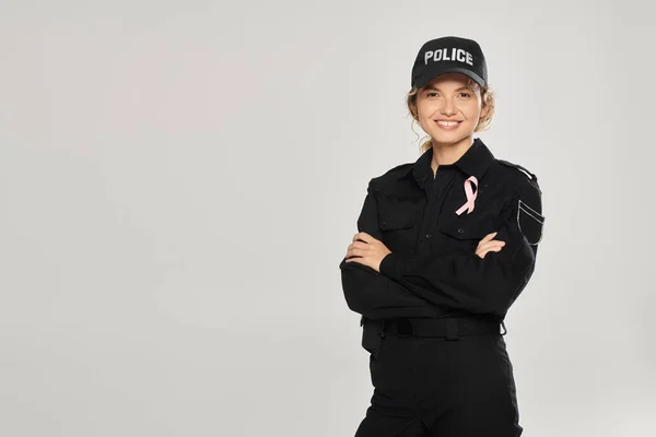 Positive policewoman with pink ribbon crossing arms isolated on grey, breast cancer concept — Stock Photo
