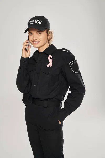 Glückliche Polizistin mit rosafarbener Schleife an Uniform, die auf dem Smartphone mit grauer Isolierung spricht, Brustkrebs — Stockfoto