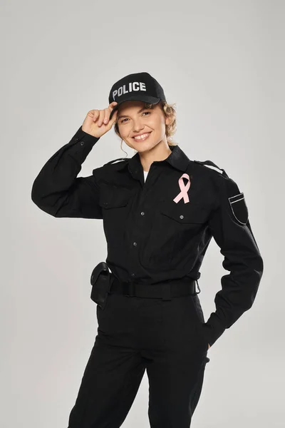 Policial sorridente com fita rosa de consciência de câncer de mama olhando para a câmera isolada em cinza — Fotografia de Stock