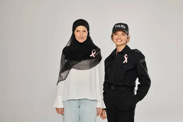 Smiling policewoman and woman in hijab with pink ribbons of breast cancer isolated on grey — Stock Photo