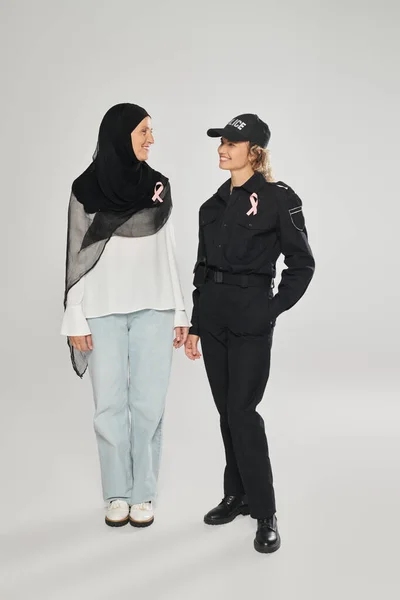 Позитивная женщина-полицейский и женщина в хиджабе с розовыми лентами рака молочной железы, стоящие на сером — стоковое фото