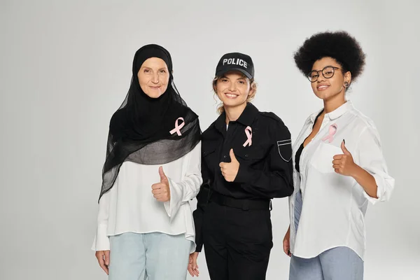 Smiling multiethnic women with pink ribbons showing like gesture isolated on grey, breast cancer — Stock Photo