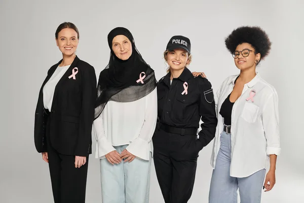 Smiling different interracial women with pink ribbons of breast cancer standing isolated on grey — Stock Photo