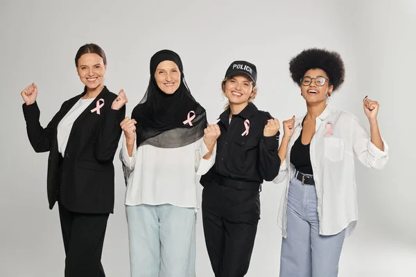 Mujeres multiétnicas excitadas y diferentes con cintas rosadas de cáncer de mama posando aisladas en gris - foto de stock