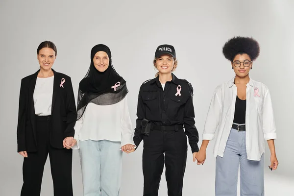 Sorridente diverse donne interrazziale con nastri rosa che tengono le mani isolate su grigio, cancro al seno — Foto stock
