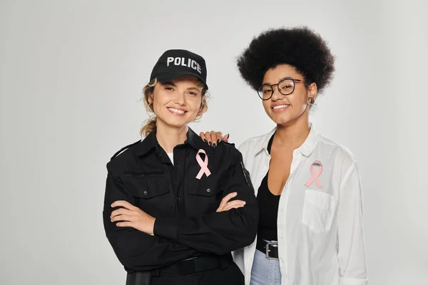 Smiling african american woman with pink ribbon hugging policewoman isolated on grey, breast cancer — Stock Photo