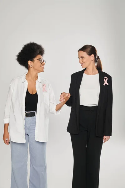 Smiling businesswoman and african american woman with pink ribbons holding hands isolated on grey — Stock Photo