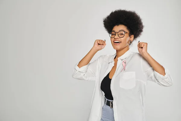 Excited african american woman with pink ribbon of breast cancer standing isolated on grey — Stock Photo