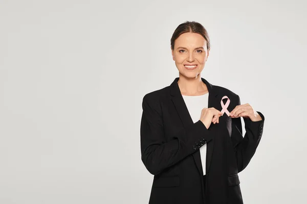 Positive businesswoman in suit holding pink ribbon of breast cancer isolated on grey — Stock Photo