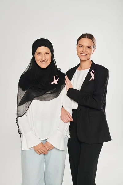 Donna d'affari sorridente che abbraccia una donna in hijab con nastro rosa isolato su grigio, mese del cancro al seno — Foto stock