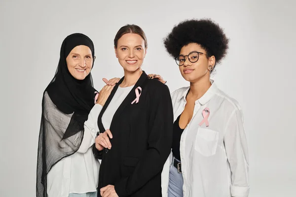 Sorridente interracial mulheres com fitas rosa de câncer de mama abraço isolado no cinza — Fotografia de Stock