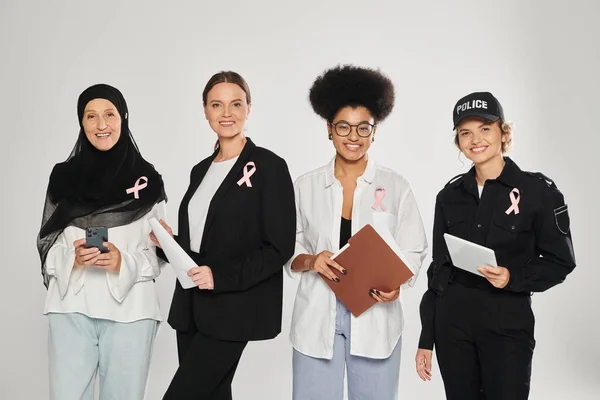 Lächelnd verschiedene interrassische Frauen mit rosa Schleifen, die Geräte und Papiere isoliert auf grau halten — Stockfoto