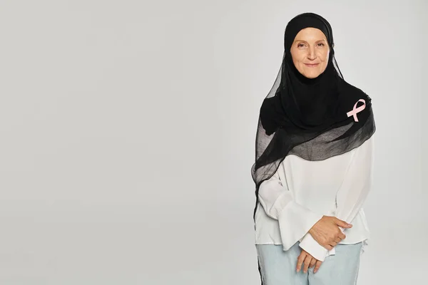 Donna allegra in hijab e nastro rosa di consapevolezza del cancro al seno in piedi isolato su grigio — Foto stock