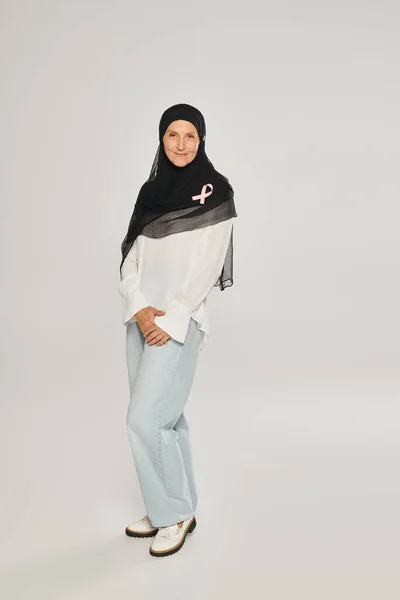 Smiling woman in hijab with pink ribbon of breast cancer awareness standing on grey background — Stock Photo