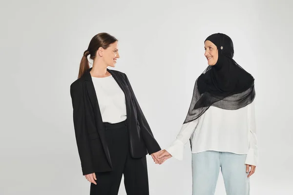 Mujer de negocios sonriente en traje y mujer en hijab tomados de las manos aislados en gris - foto de stock