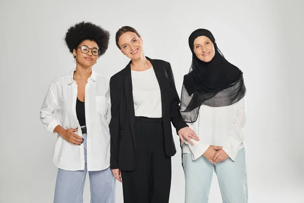 Smiling multiethnic women looking at camera while posing together isolated on grey — Stock Photo