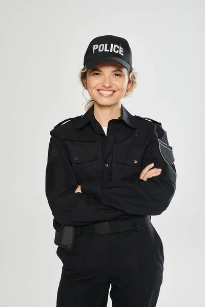 Cheerful policewoman in uniform crossing arms and posing isolated on grey — Stock Photo