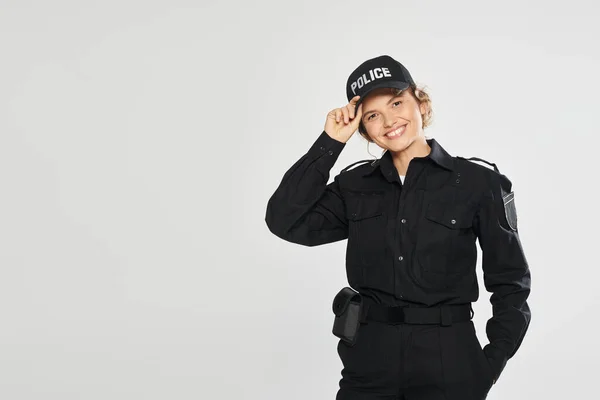 Cheerful policewoman in uniform touching cap and looking at camera isolated on grey — Stock Photo