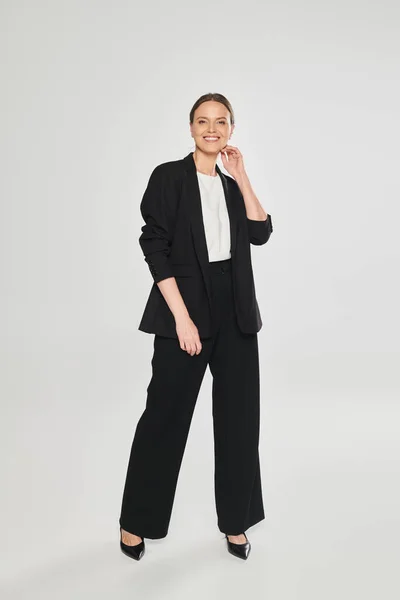 Volle Länge der fröhlichen Frau in High Heels und Anzug auf grauem Hintergrund stehend — Stockfoto