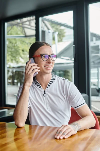 Молодой человек с хвостом и очками разговаривает по телефону за столом со стеклом на заднем плане, коворкинг — стоковое фото