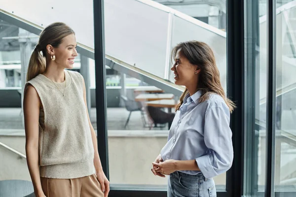 Smiling female colleagues in smart casual attire looking at each other with window backdrop — Stock Photo