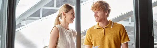 Jóvenes compañeros de trabajo en ropa inteligente sonriendo y sosteniendo papeles con telón de fondo de vidrio, concepto de coworking - foto de stock