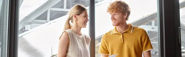 Jóvenes compañeros de trabajo en ropa inteligente sonriendo y sosteniendo papeles con telón de fondo de vidrio, concepto de coworking — Stock Photo