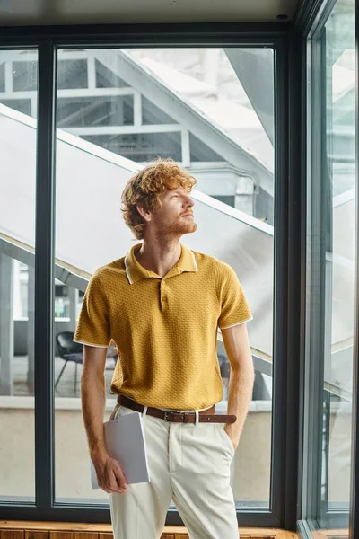 Jovem homem ruivo em camisa amarela casual inteligente olhando para longe com o pano de fundo da janela, coworking — Fotografia de Stock