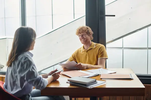 Colegas felizes sentados à mesa e sorrindo um para o outro segurando telefone e papéis de trabalho, co-trabalhando — Stock Photo