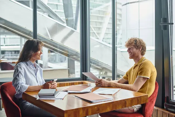 Dos colegas sonrientes en ropa elegante con documentos en la mesa mirándose, coworking - foto de stock