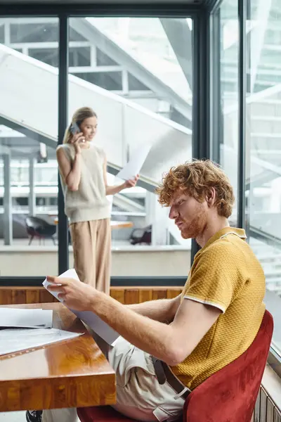 Homem de cabelo vermelho olhando para documentos e seu colega falando no telefone no pano de fundo, conceito de co-working — Fotografia de Stock