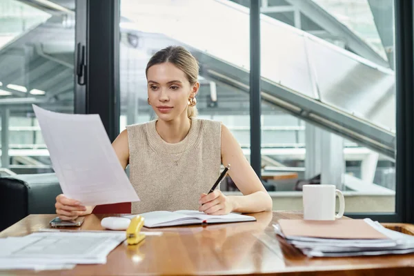 Молодая сотрудница в деловой повседневной одежде сидит за столом и смотрит на свои документы, коворкинг — стоковое фото