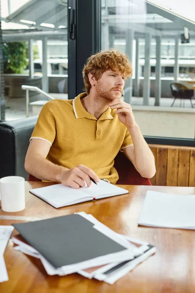 Rothaariger junger Mann, der an seinen Papieren arbeitet und nachdenklich wegschaut, Coworking-Konzept — Stockfoto
