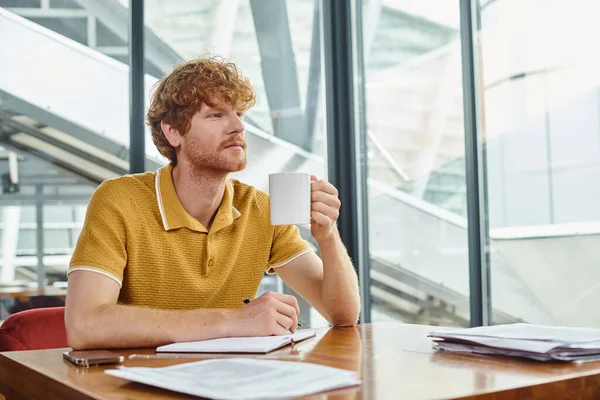 Nachdenklicher junger Mann mit roten Haaren, der an seinen Dokumenten arbeitet und Teetasse in der Hand hält, Coworking-Konzept — Stockfoto