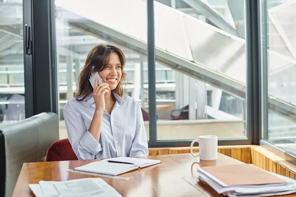 Cheerful young employee in business casual outfit sitting and talking on phone, coworking concept — Stock Photo