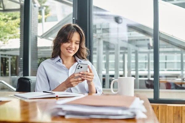 Felice donna bruna in abbigliamento intelligente che lavora a tavola e guarda smartphone, concetto di coworking — Foto stock