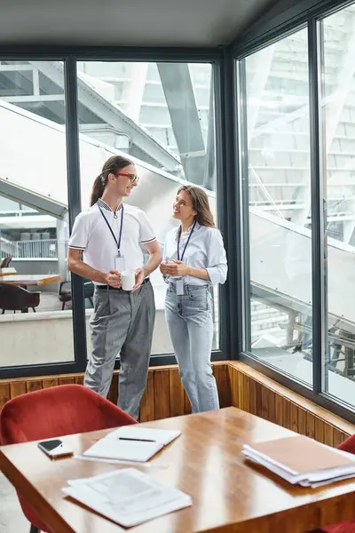Dos colegas alegres de pie detrás de la mesa con papelería y mirándose, coworking - foto de stock