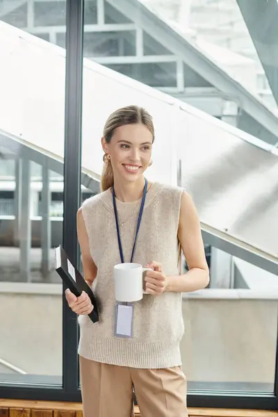 Giovane donna bionda in smart wear con tazza di tè e taccuino sorridente e distogliendo lo sguardo, coworking — Foto stock
