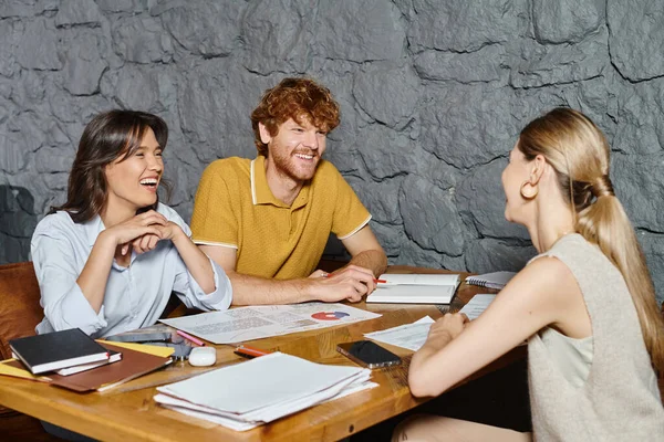 Tres compañeros alegres riendo y mirándose mientras trabajan en papeles, coworking - foto de stock
