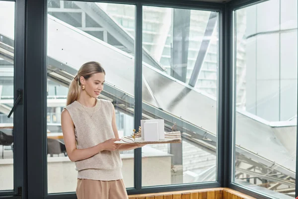 Young attractive employee holding scale model of building with glass on background, design bureau — Stock Photo