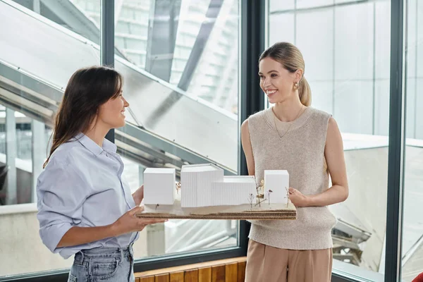 Команда из двух молодых женщин, держащих модель здания и смотрящих друг на друга, конструкторское бюро — стоковое фото