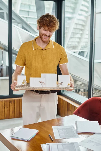 Red haired man smiling and holding architecture model with glass on background, design bureau — Stock Photo