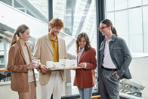 Young elegant team looking at scale model and holding tea cup with glass backdrop, design bureau — Stock Photo