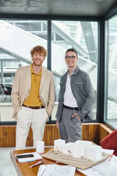 Zwei männliche Mitarbeiter posieren am Tisch mit maßstabsgetreuem Modell und schauen in die Kamera, Designbüro — Stockfoto