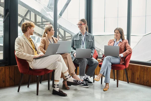 Four cheerful colleagues looking at each other and working on laptops with glass backdrop, coworking — Stock Photo